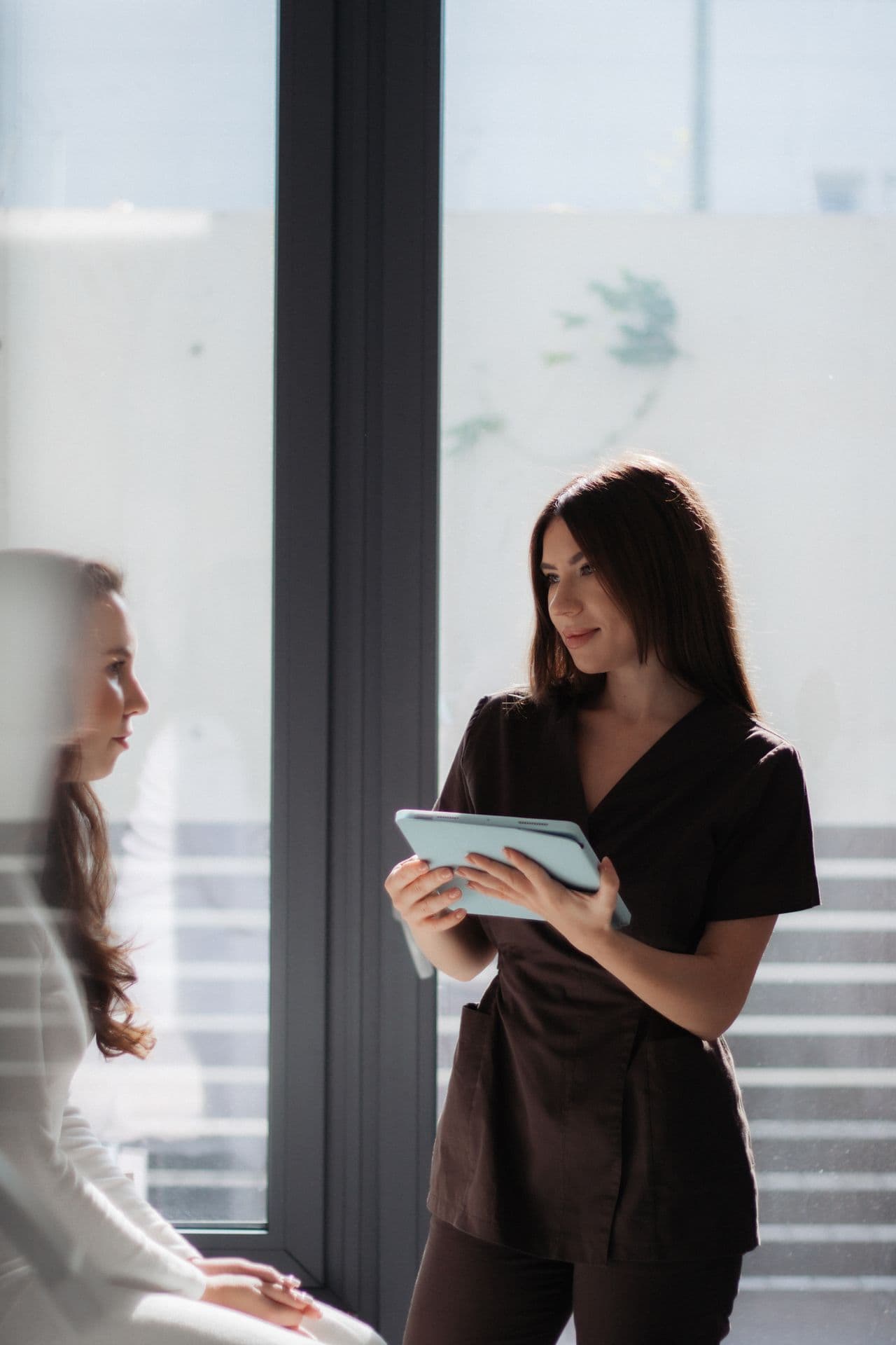 A practitioner holding a tablet while in conversation with a client in a calm, sunlit aesthetic clinic